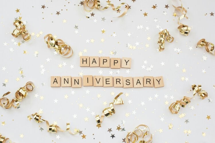 Scrabble tiles spell "HAPPY ANNIVERSARY" surrounded by gold ribbons and star confetti on a white background.