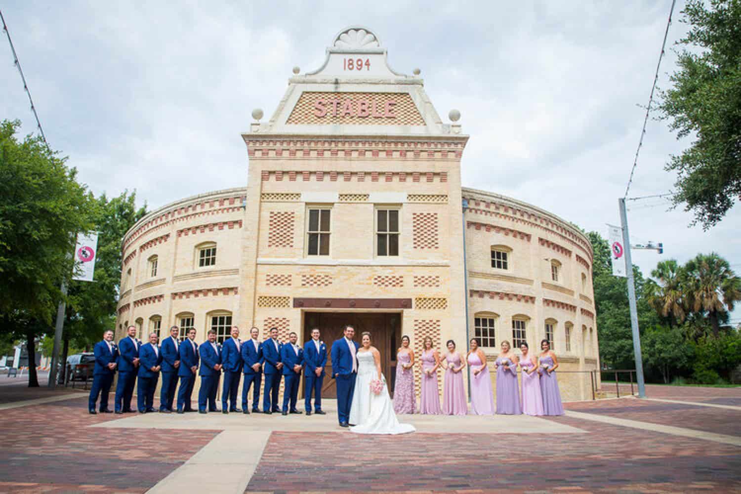 Wedding party poses in front of a historic brick stable, with the bride, groom, bridesmaids in pink dresses, and groomsmen in blue suits.
