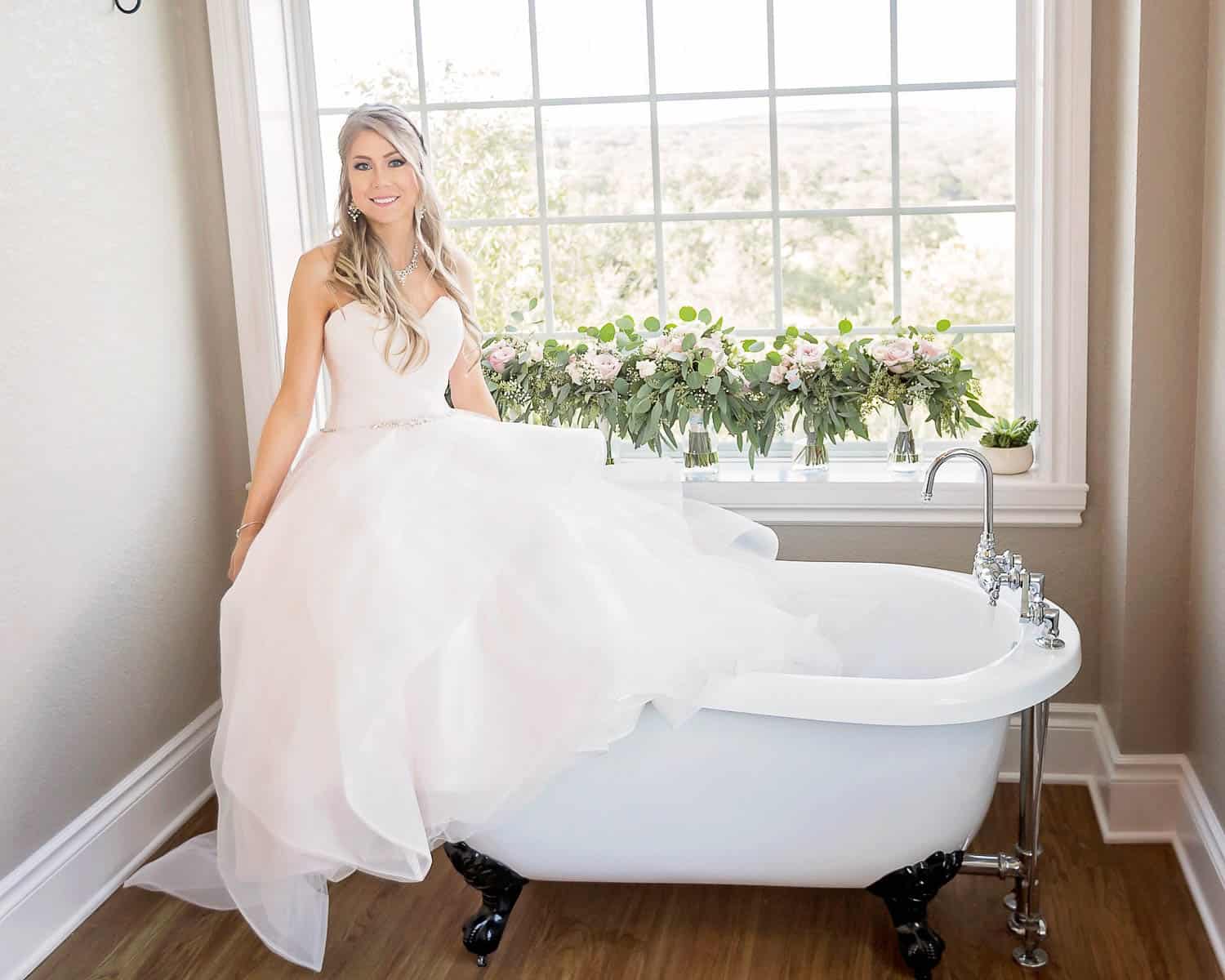 A woman in a white gown sits on the edge of a vintage bathtub in a bright room with a large window and floral decorations.