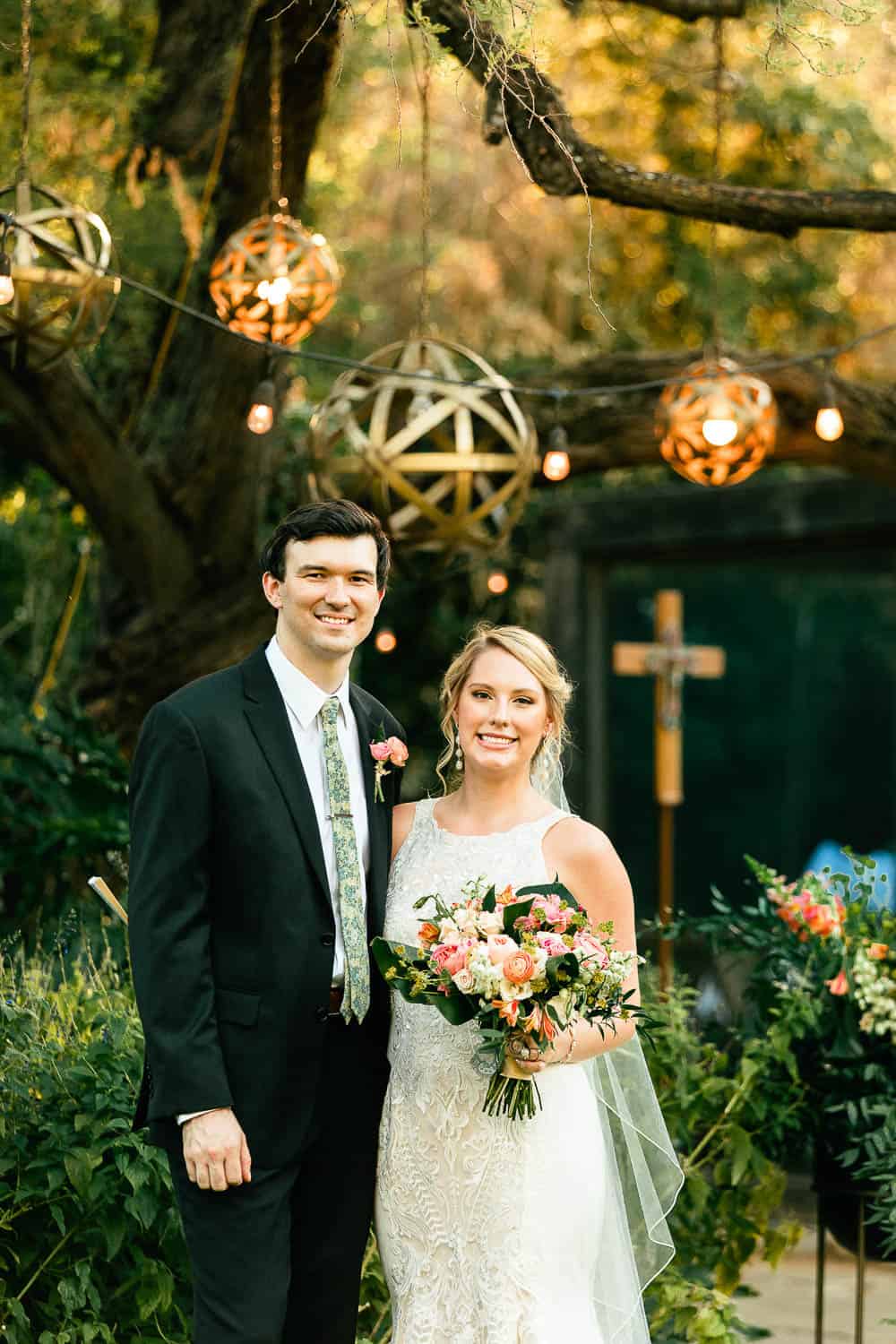 A couple in formal attire poses outdoors. The woman holds a bouquet, and decorative lights hang above them.