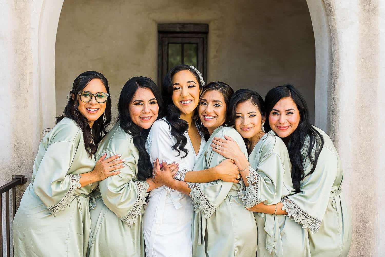Six women in matching robes, smiling and embracing each other in front of an arched doorway.