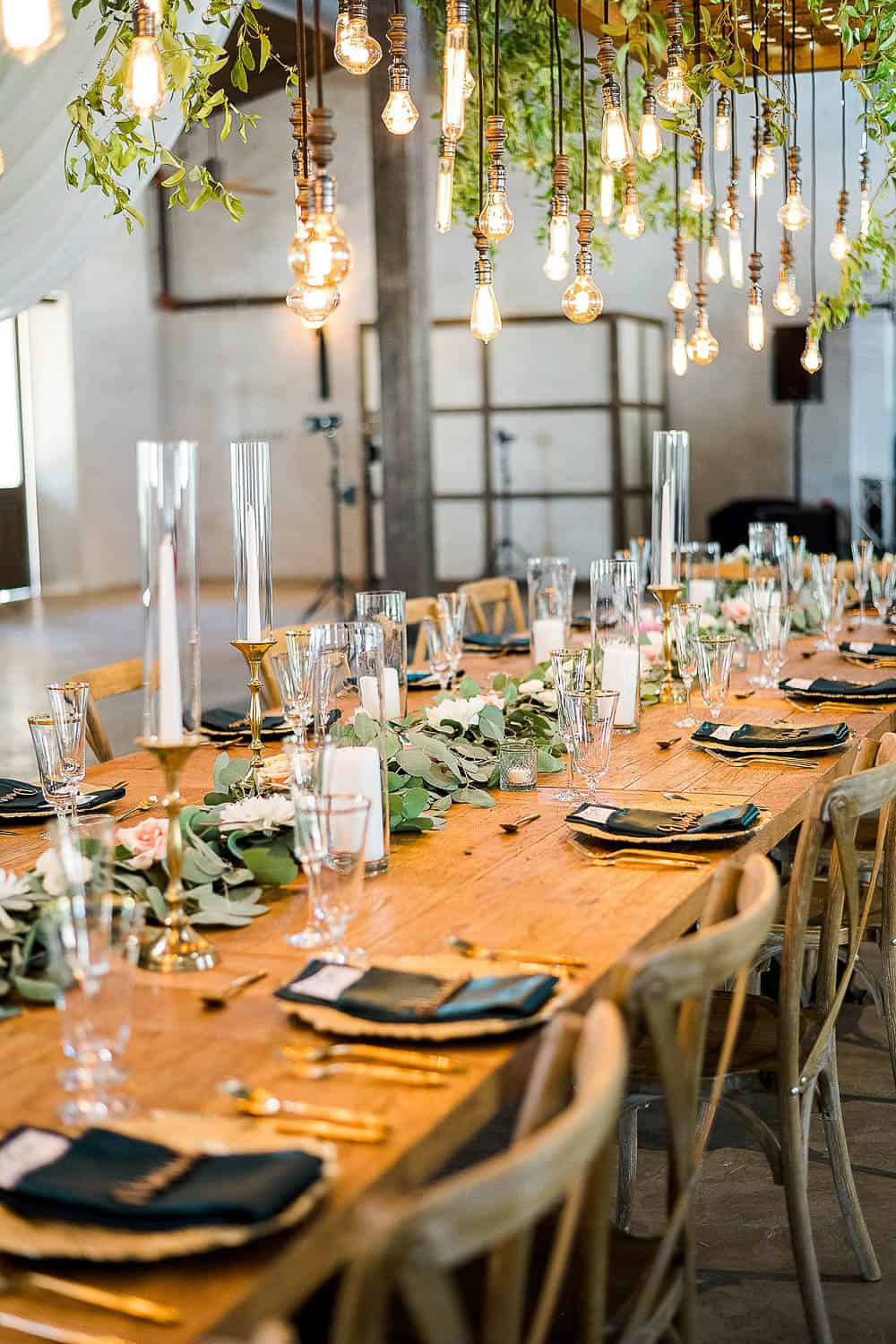 A long wooden dining table is set with green napkins, glasses, and gold cutlery. Greenery and hanging bulbs decorate the area above the table.