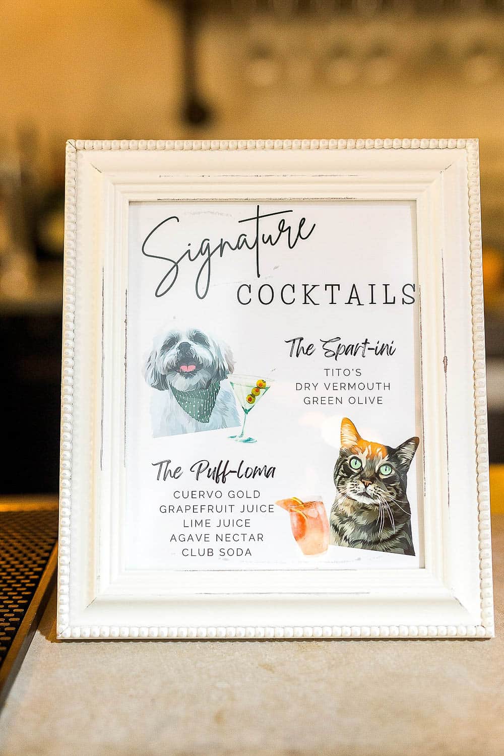Framed cocktail menu featuring "The Spart-ini" with a dog illustration and "The Puff-Loma" with a cat illustration, listing ingredients for each drink.