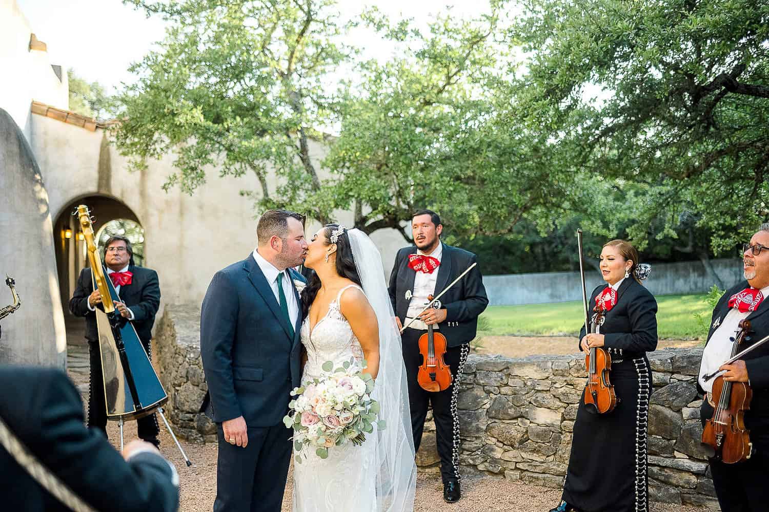 Bride and groom kiss outdoors, surrounded by a mariachi band in black suits with red ties. The bride holds a bouquet and wears a veil; the groom is in a blue suit. Stone wall and trees in the background.