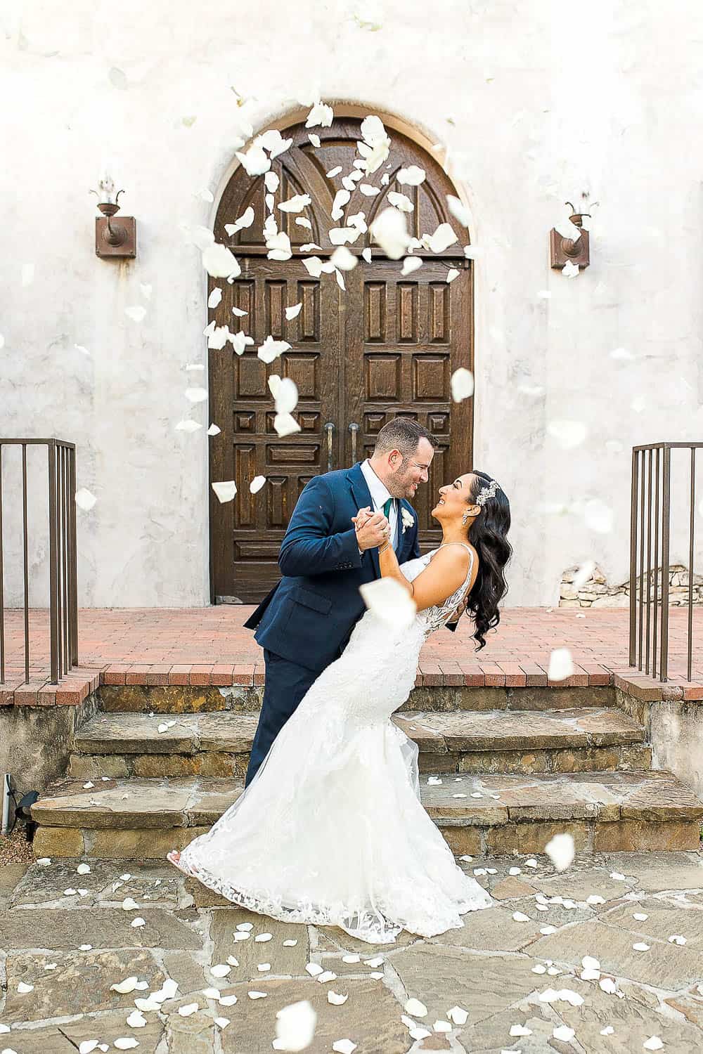 A couple in wedding attire dances joyfully on stone steps, surrounded by falling white rose petals, in front of a large wooden door.