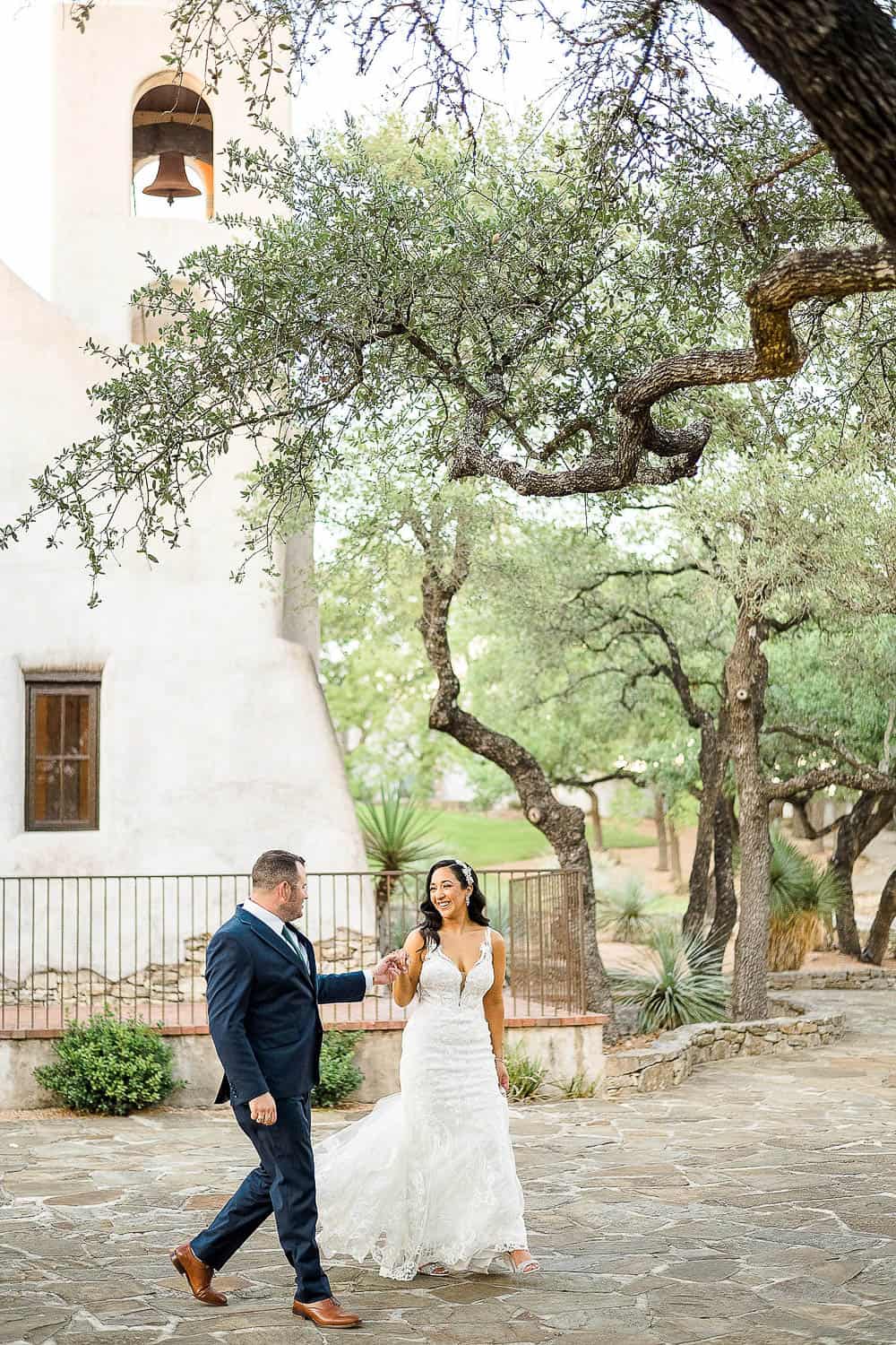 A couple in formal attire walks joyfully hand in hand on a stone path near a bell tower and trees.