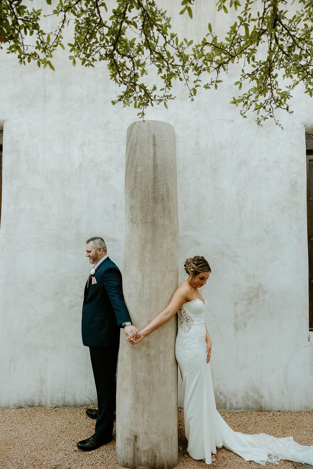 A bride and groom stand back-to-back against a tall stone column, holding hands, in front of a textured wall with a branch above them.