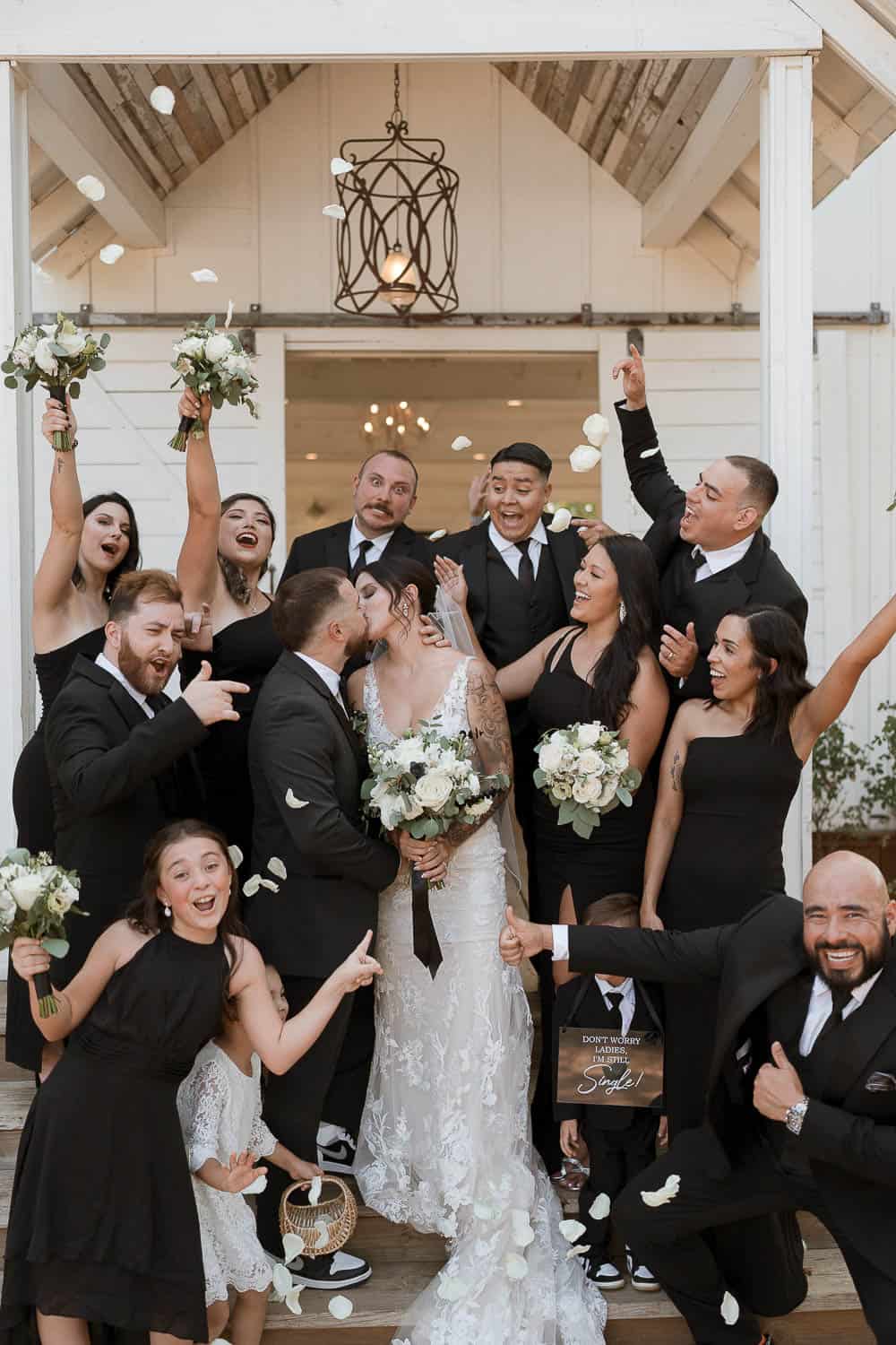 Wedding party celebrating on a porch as the bride and groom kiss in the center. Guests in formal attire cheer and throw flower petals.