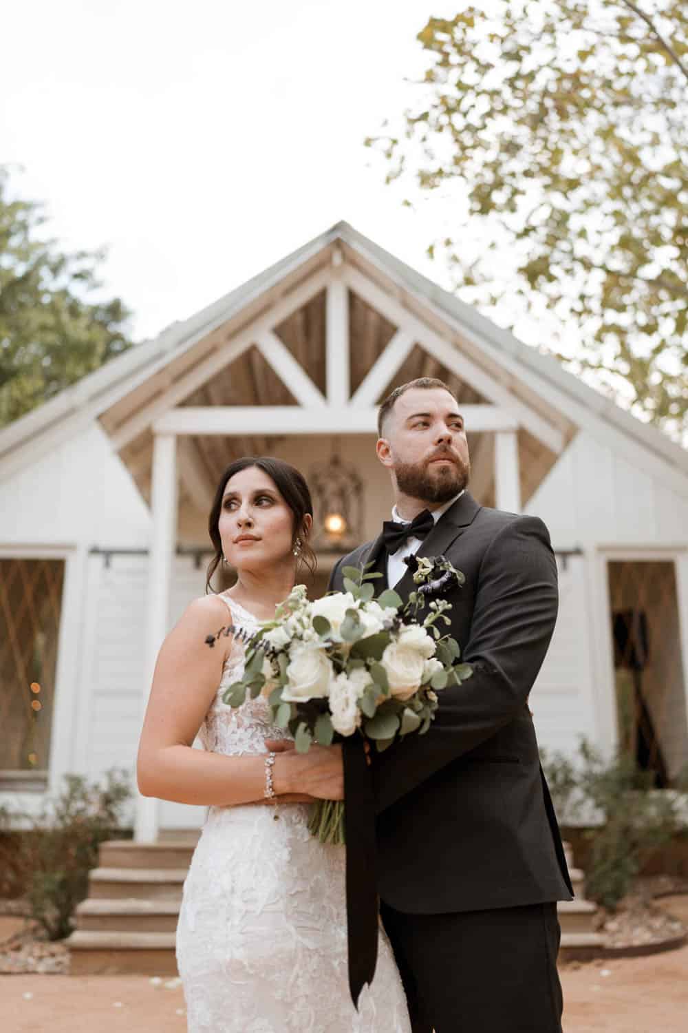 A bride and groom stand in front of a small chapel, holding a bouquet. They look in opposite directions, dressed in formal wedding attire.