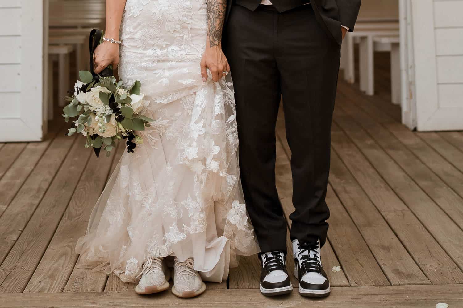 Couple dressed in wedding attire; the bride in a lace gown with sneakers, holding a bouquet, and the groom in a suit with sneakers, standing on a wooden deck.