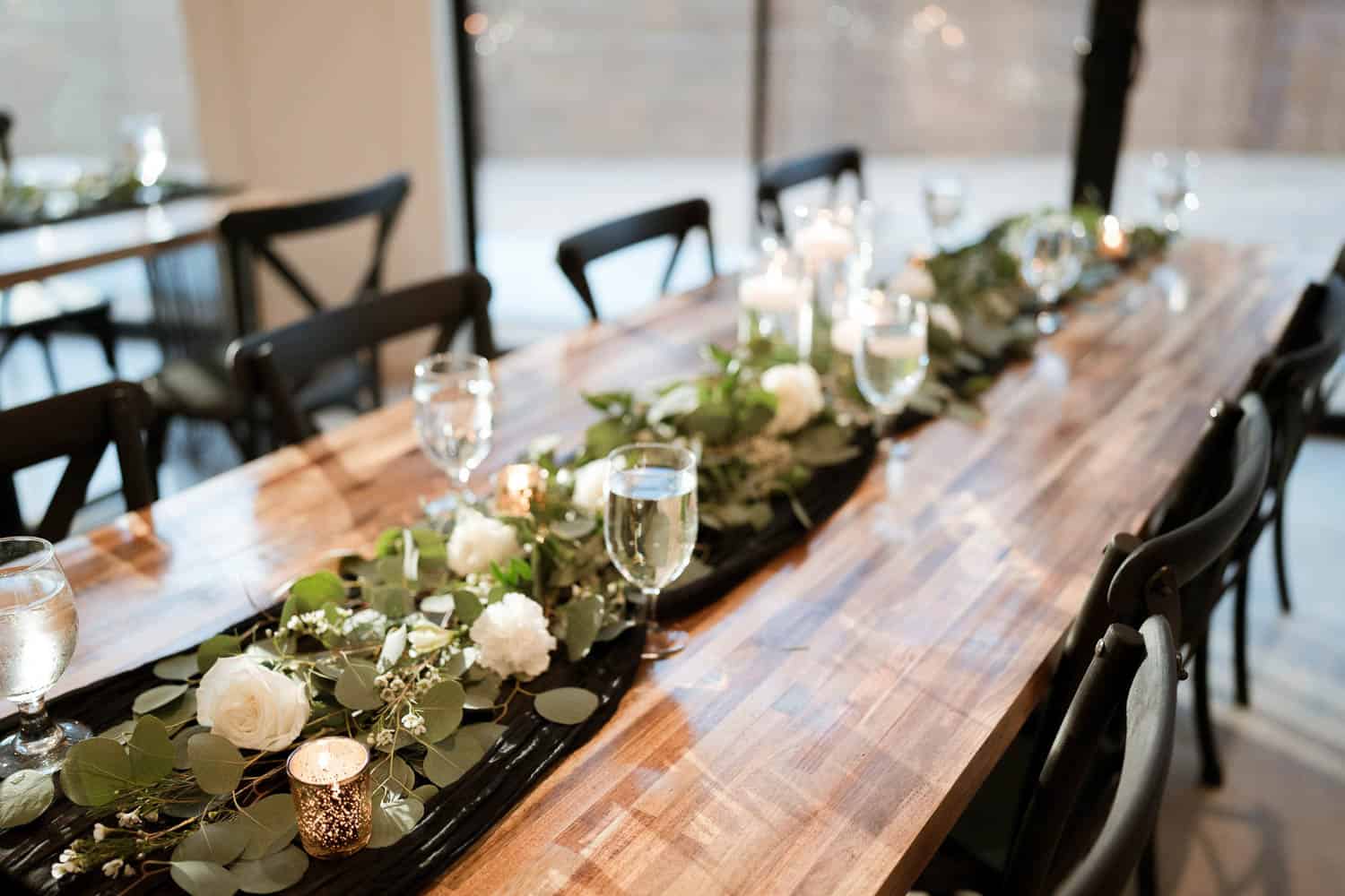 A wooden table is set with green and white floral centerpieces, candles, and glasses of water, with black chairs on either side.