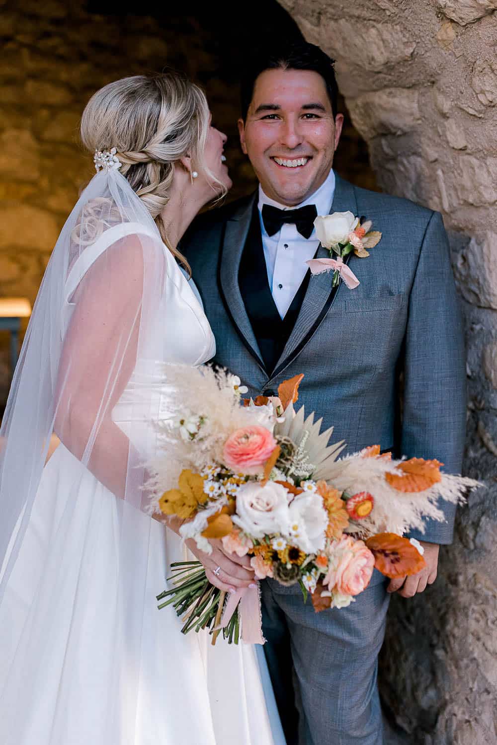 Bride in white dress and veil whispers to smiling groom in gray suit with bow tie. She holds a bouquet of roses and dried leaves. Stone wall in the background.