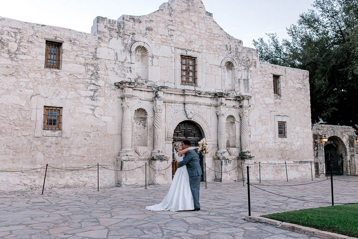 A bride and groom embrace in front of a historic stone building with arched windows and a carved entrance.