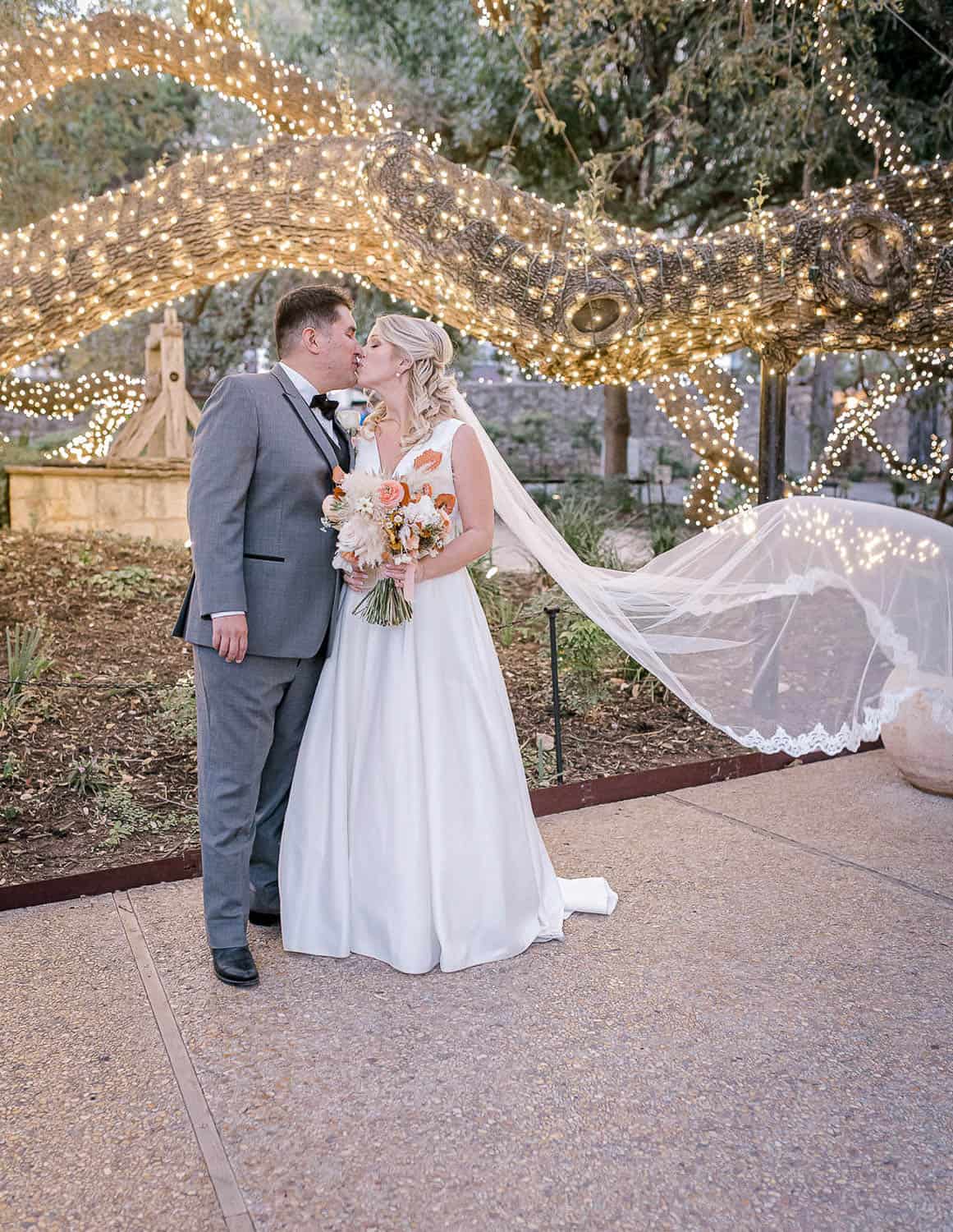 A bride and groom share a kiss under a tree decorated with string lights. The bride holds a bouquet and her veil flows behind her.