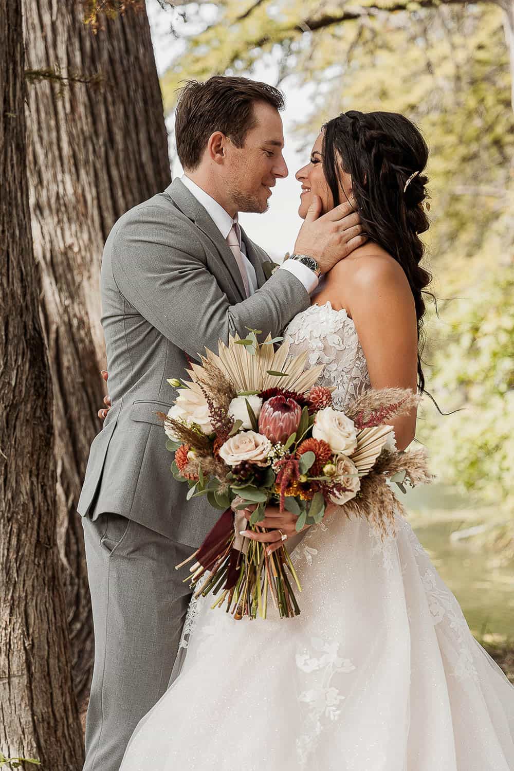 Couple embraces in a forest. The man wears a gray suit, and the woman in a white wedding dress holds a bouquet. They smile and look at each other.