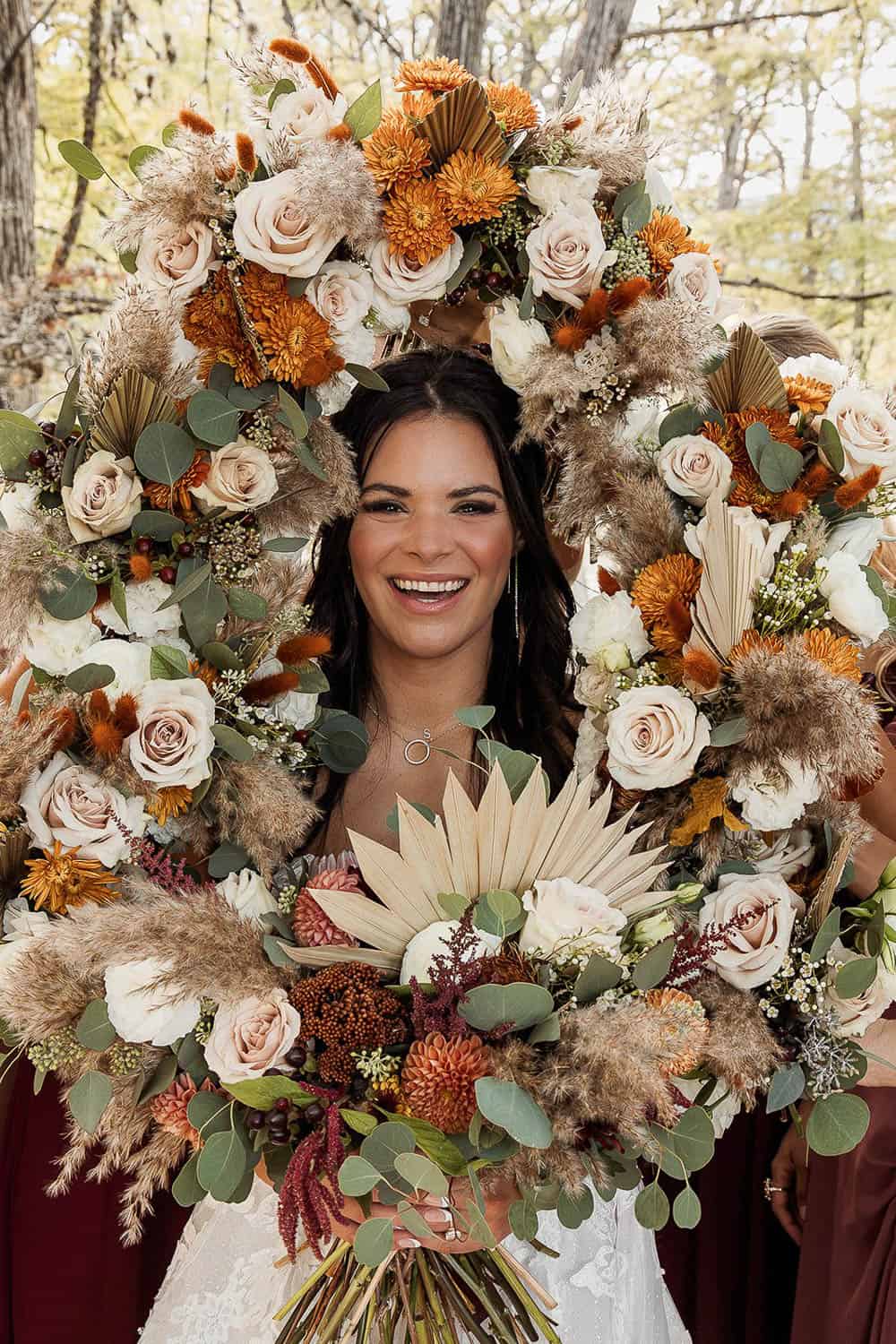 A woman smiles, holding a large floral wreath made of white roses, orange flowers, and green leaves. She wears a white dress and has long, dark hair.