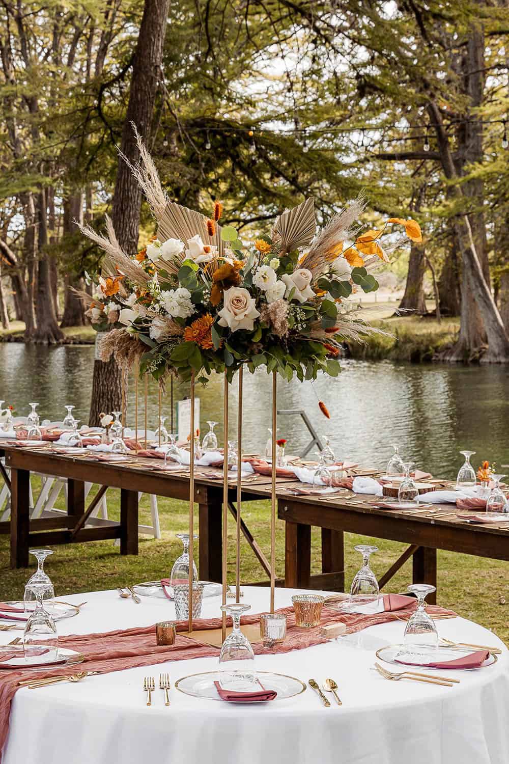 Outdoor wedding table by a lake with a tall floral and greenery centerpiece. Glassware and cutlery are arranged on white tablecloths.