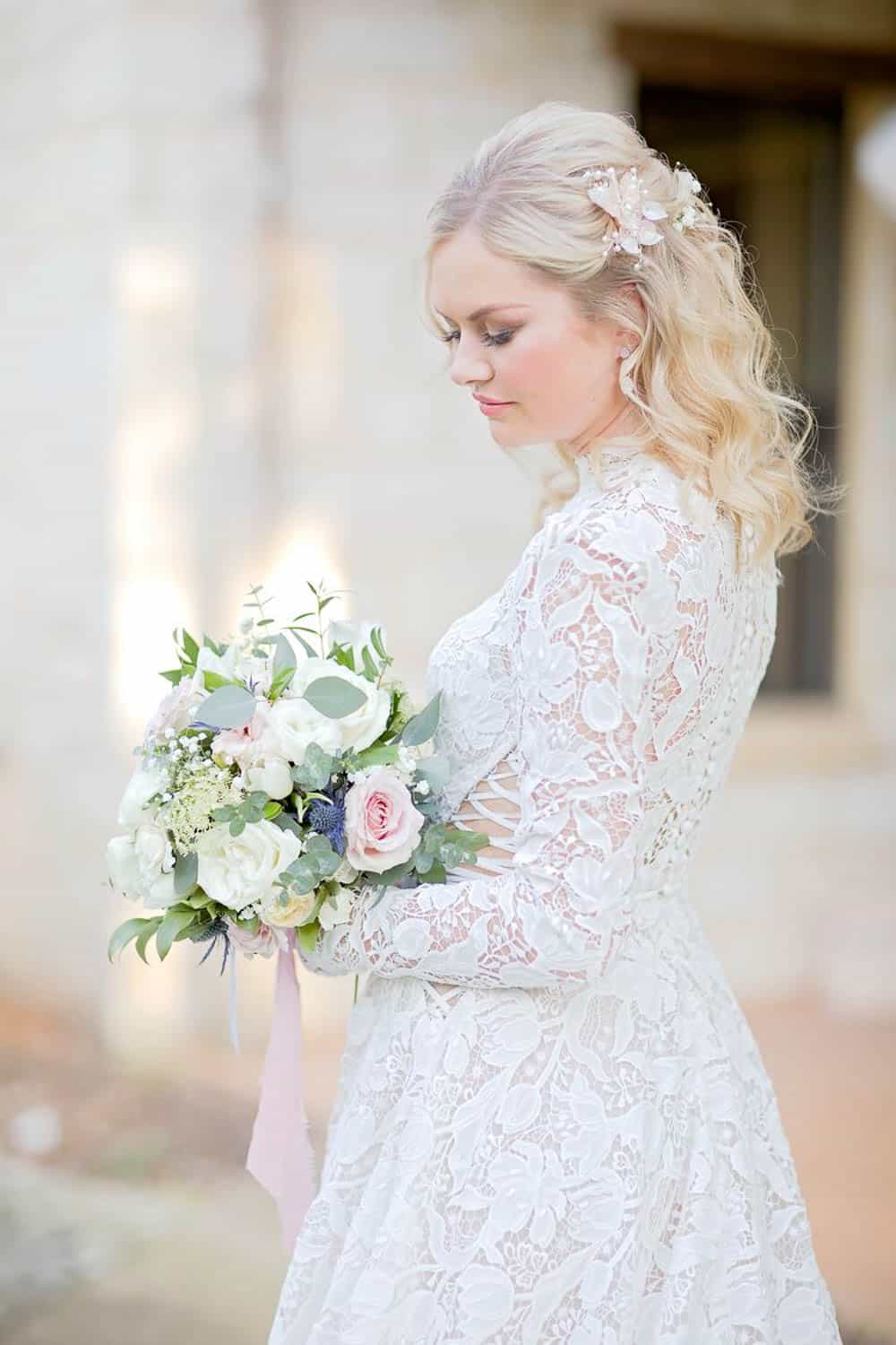 Bride in a lace wedding dress holding a bouquet of white and pink flowers, standing outdoors.