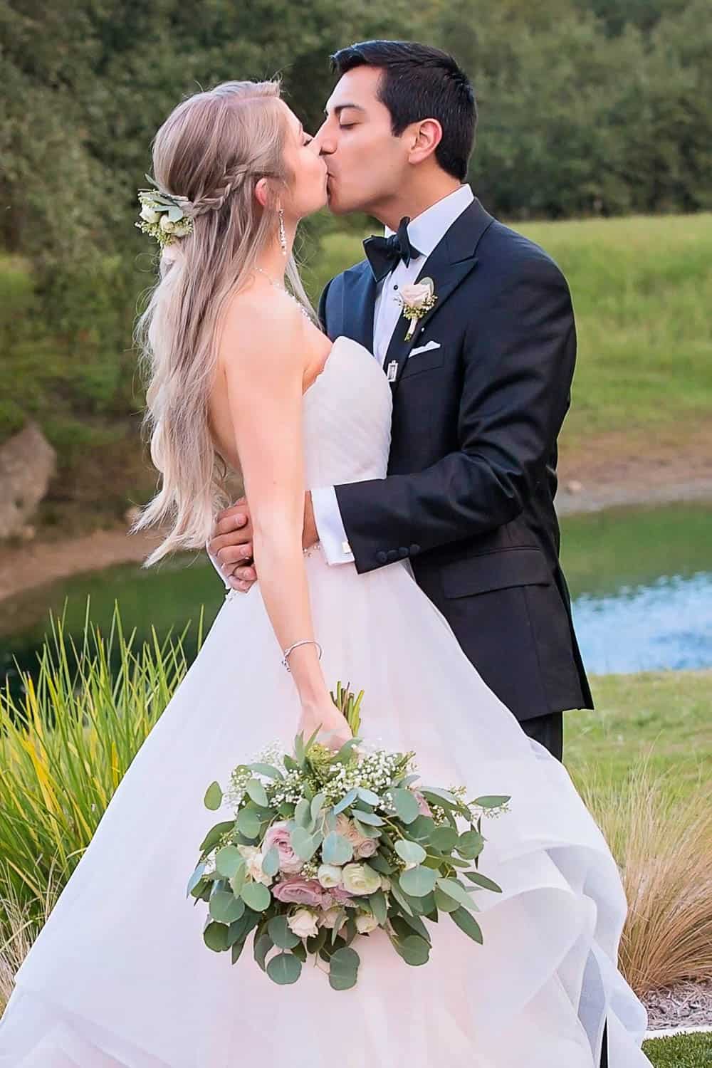 Bride and groom kiss outdoors. The bride holds a bouquet in a white gown, and the groom wears a black suit. Greenery and a pond are in the background.