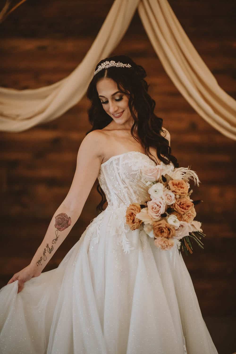 Bride in a strapless dress with a tiara, long wavy hair, and a tattoo on her arm holds a rose bouquet. Draped fabric and wooden paneling in the background.