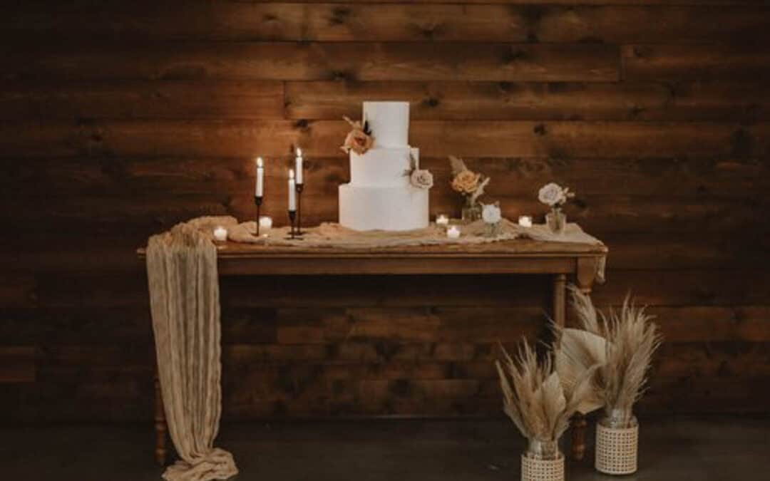 A two-tiered white cake on a wooden table with candles, flowers, and a beige cloth; vases with dried plants are on the floor in front of a wooden wall.