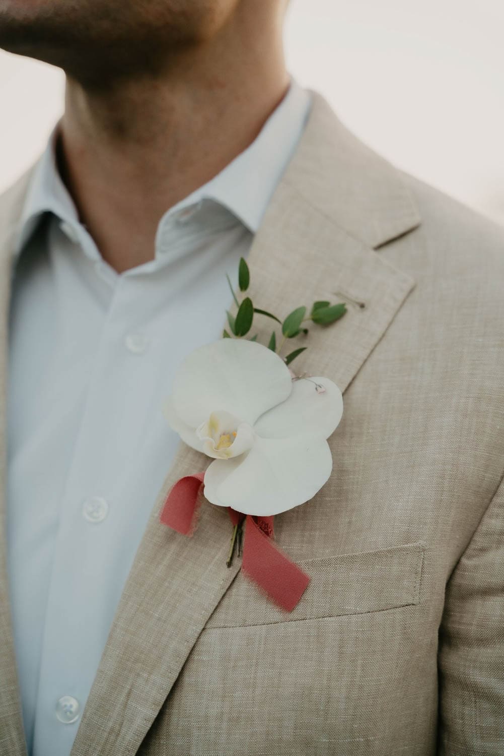 Man in a beige suit with a white orchid boutonniere and pink ribbon on the lapel, standing outdoors.