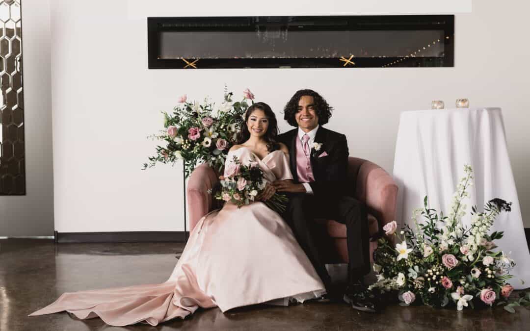 A couple sits on a pink couch with floral arrangements around them. The woman wears a pink gown, and the man wears a black suit with a pink tie.