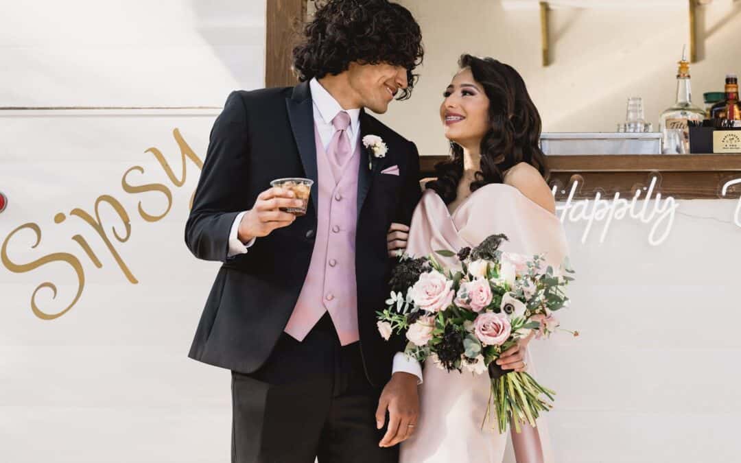 Couple in front of a bar: the man in a black suit with a pink vest holds a drink, and the woman in a pink dress holds a bouquet as they smile at each other.