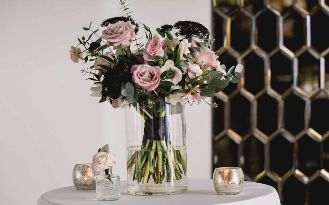 A vase filled with pink and white flowers arranged with greenery sits on a table next to two small glass candle holders.