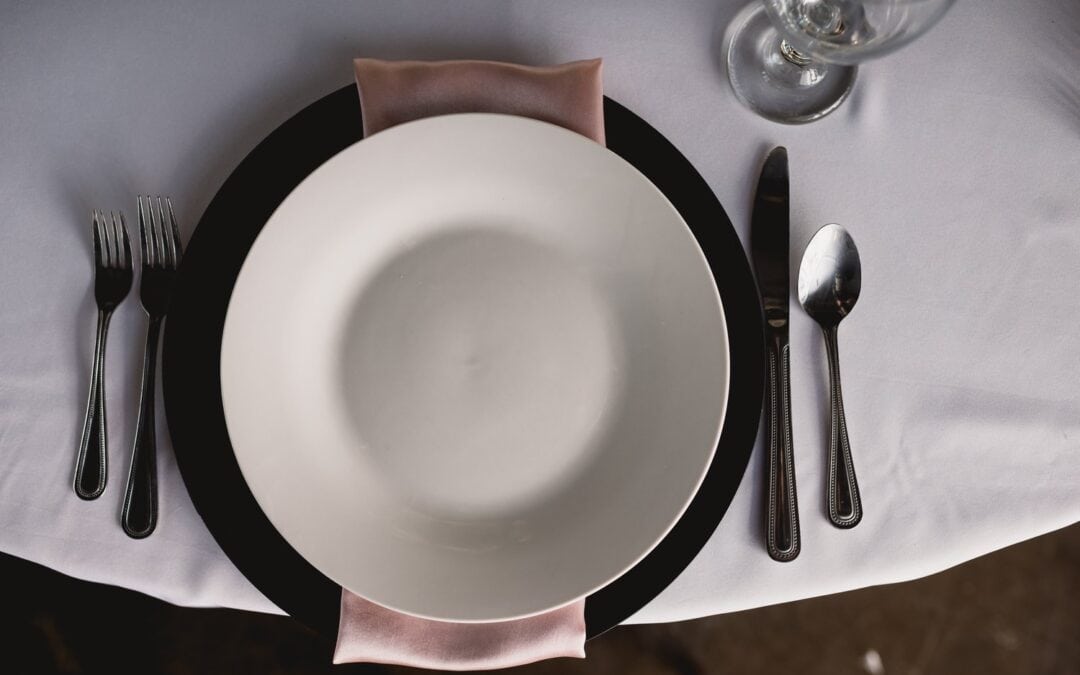 A table setting with a white plate on a black charger, pink napkin, silverware, and a wine glass on a white tablecloth.