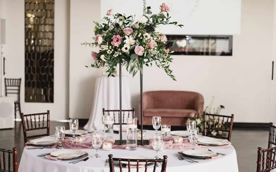 Wedding table with a tall floral centerpiece of pink and white flowers, glassware, plates, candles on a white tablecloth, and brown chairs in a bright room.