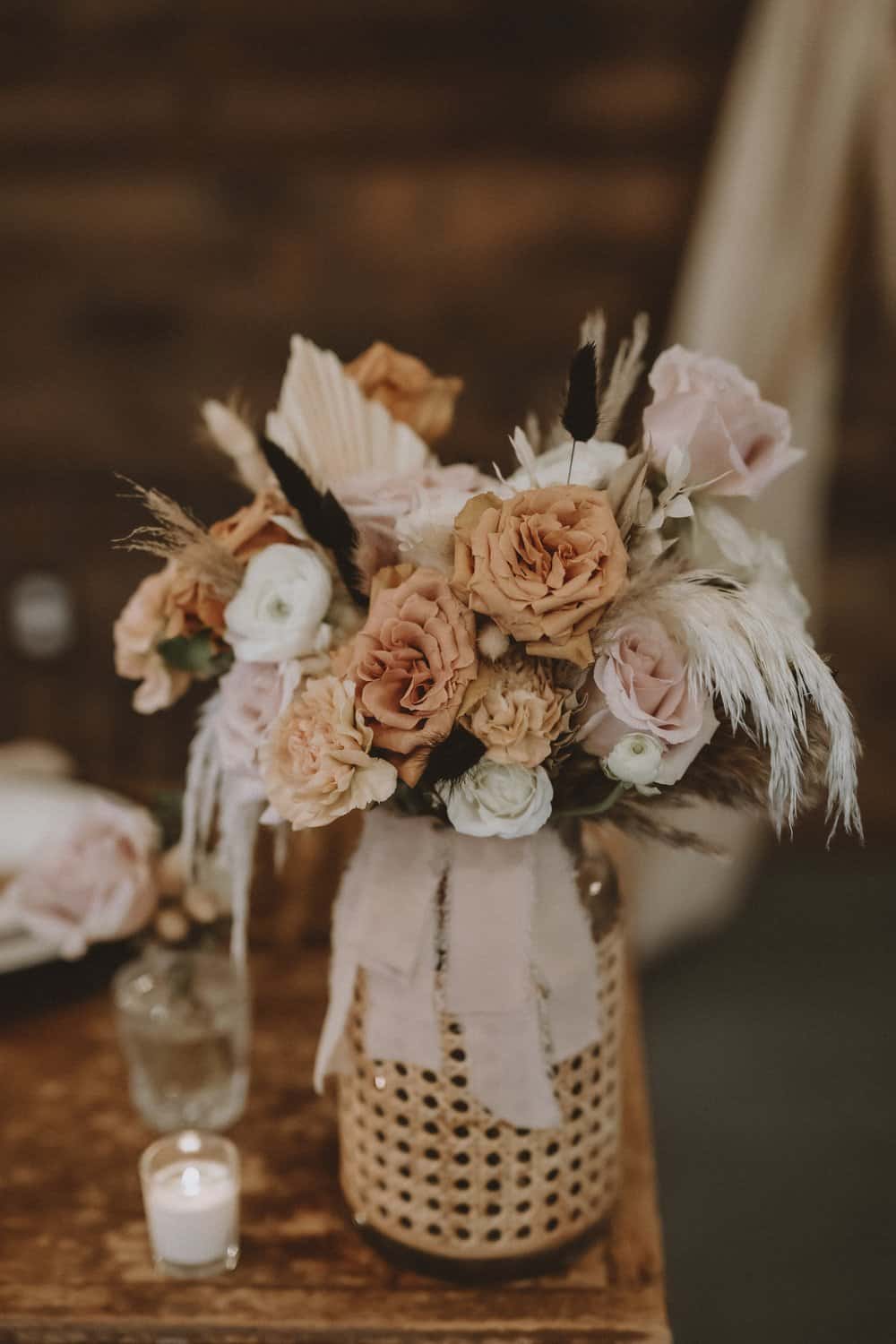 A rustic floral arrangement featuring pink, peach, and white flowers with pampas grass in a woven vase. A small candle is placed nearby on a wooden table.