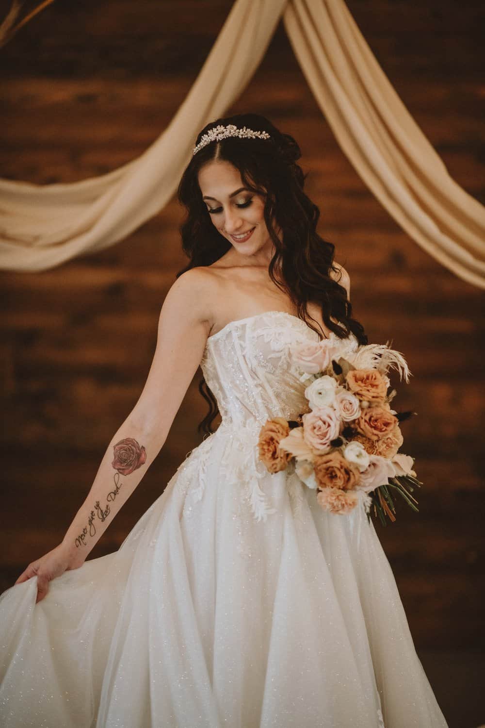 A bride in a strapless white gown holds a bouquet of roses. Draped fabric and a wooden wall create the backdrop.