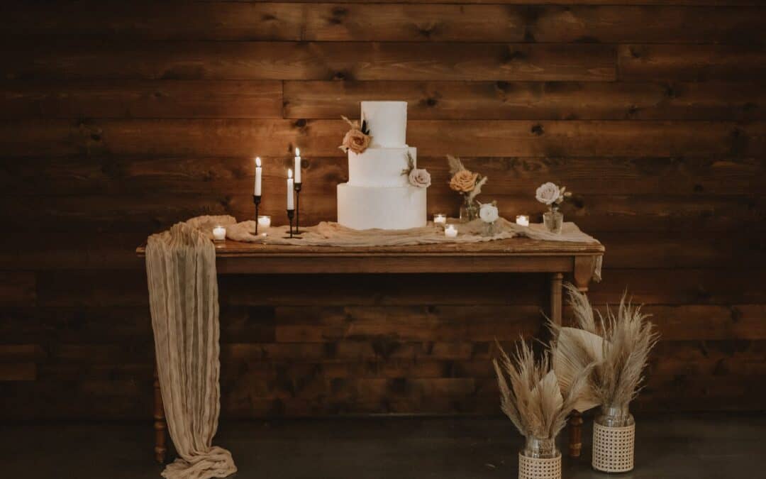 A rustic wedding cake setup on a wooden table with candles, flowers, and pampas grass decorations against a wooden backdrop.