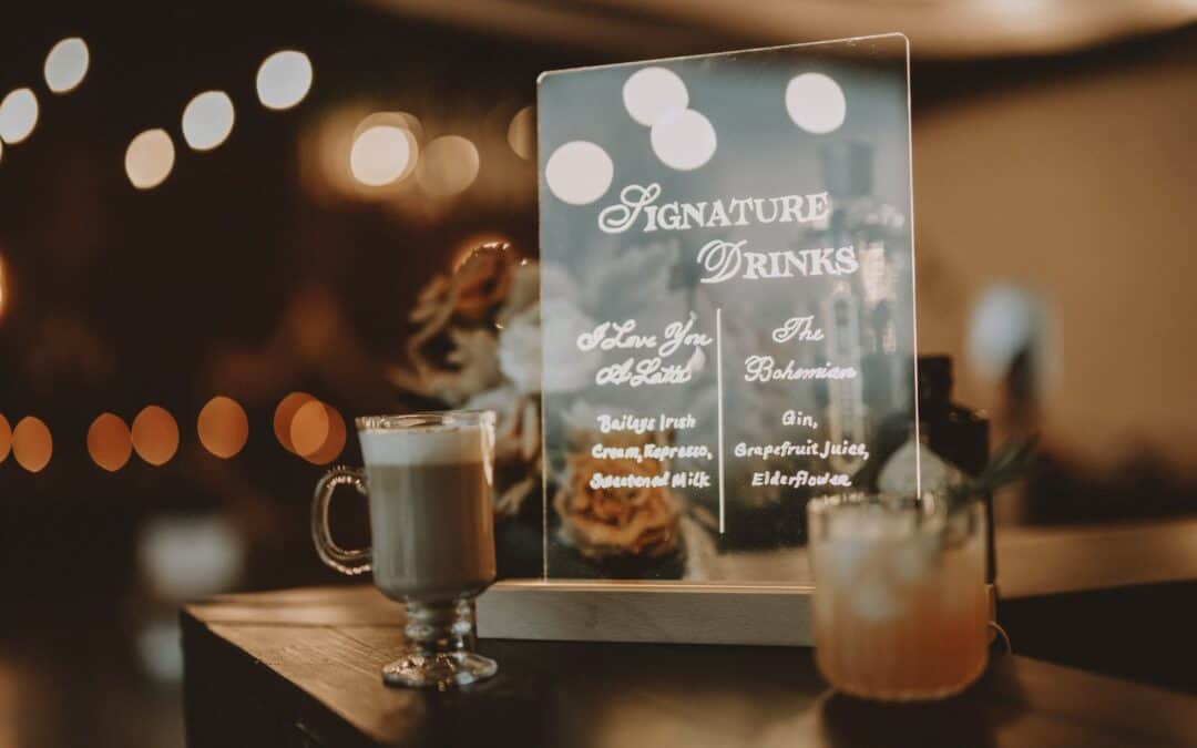 A dimly lit counter displays a sign with two signature drinks listed. A latte in a glass mug and a cocktail in a tumbler are in front of it.