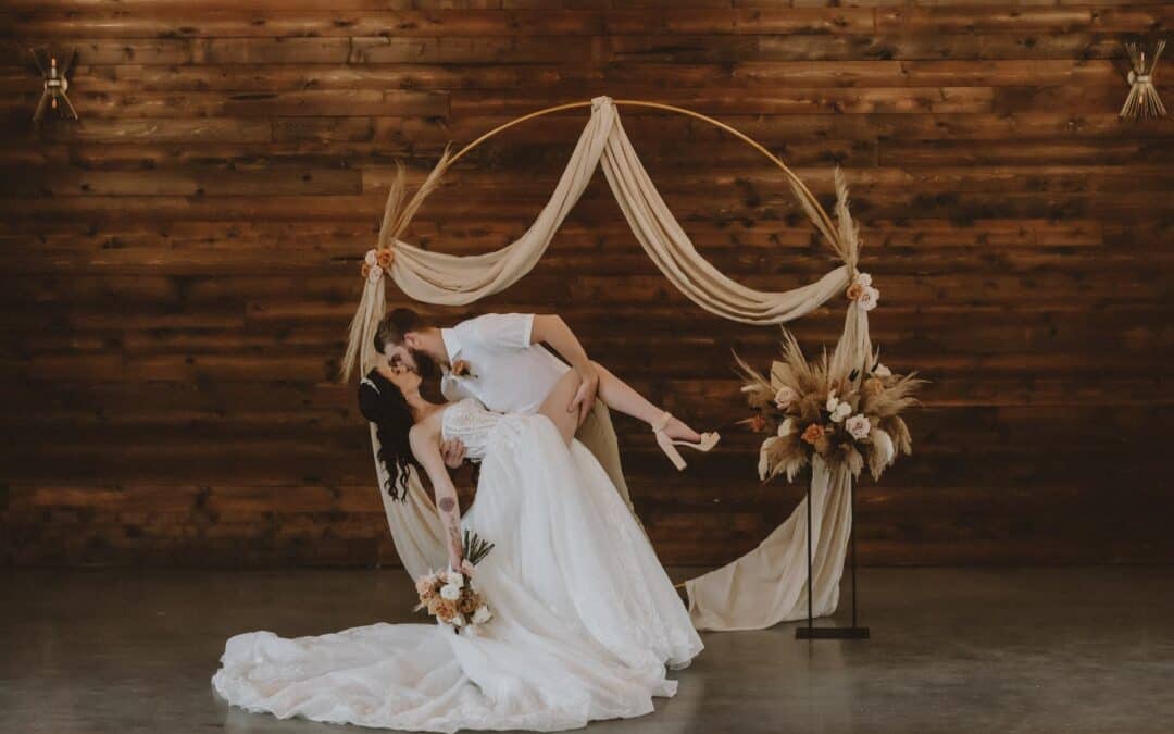 Groom dips a bride in front of a wooden backdrop with a circular arch draped in fabric and flowers.