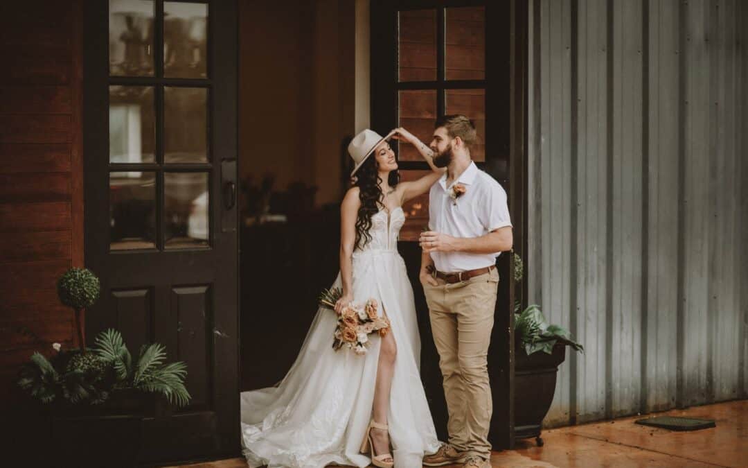 Bride in a white dress and cowboy hat stands with a man in casual attire. They're positioned in front of a rustic building, with her holding a bouquet.