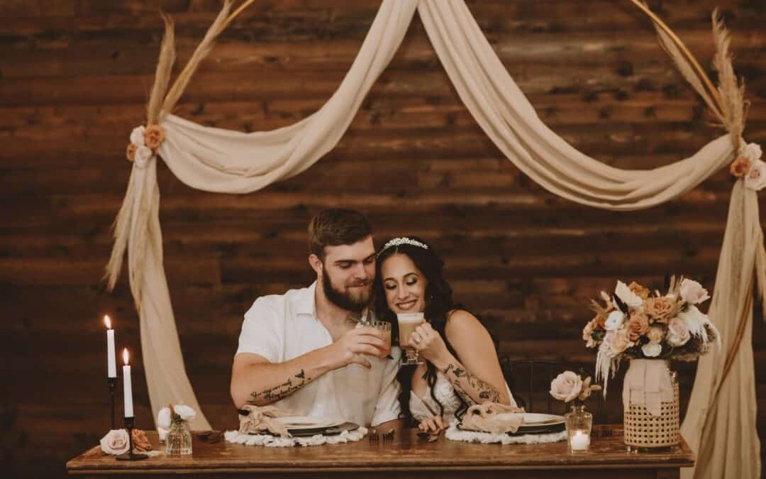 A bride and groom toasts at a decorated table with rustic flowers and candles, under a draped arch against a wooden backdrop.