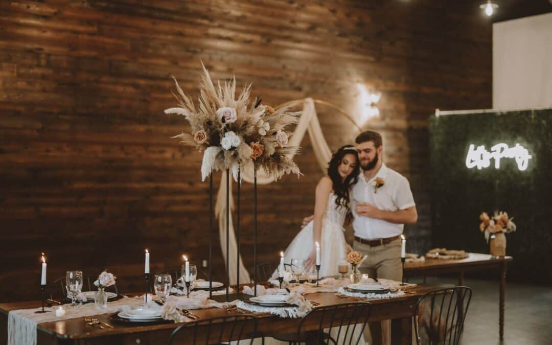 Couple stands by a wooden table set with floral centerpieces, lit candles, in a warmly lit room with dark wooden walls and a neon sign.