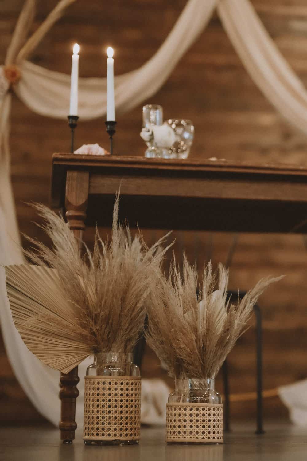 A rustic wedding table setting with two lit candles, glassware, and dried pampas grass in woven vases on the floor, against a wooden backdrop with draped fabric.