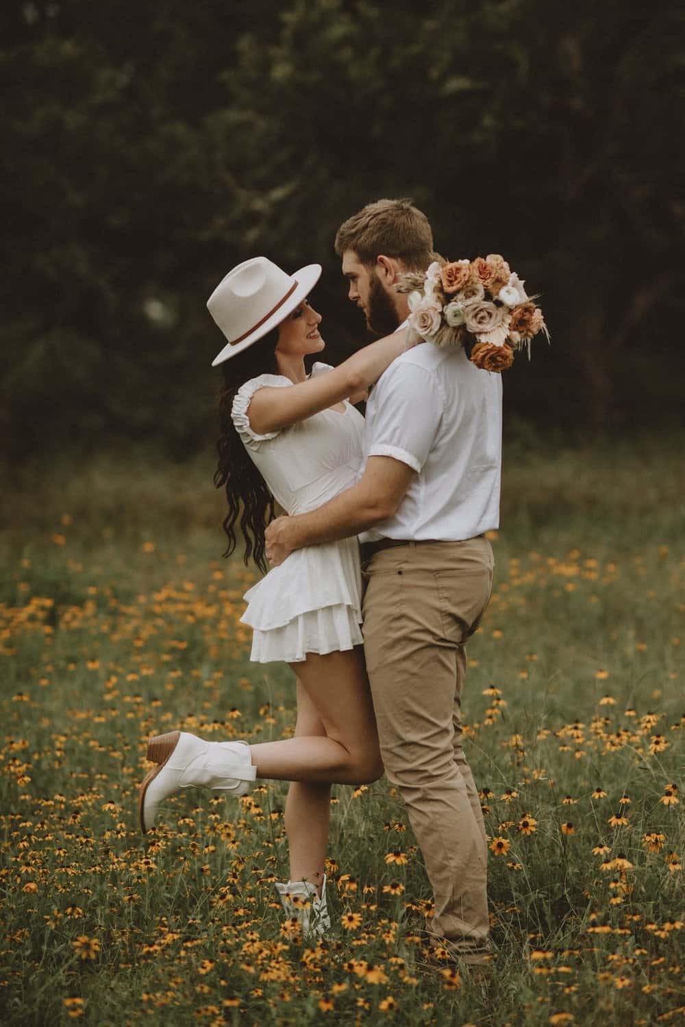 Couple embracing in a field of yellow flowers; the woman in a white dress and hat holds a white and orange bouquet, and the man wears a white shirt and beige pants.