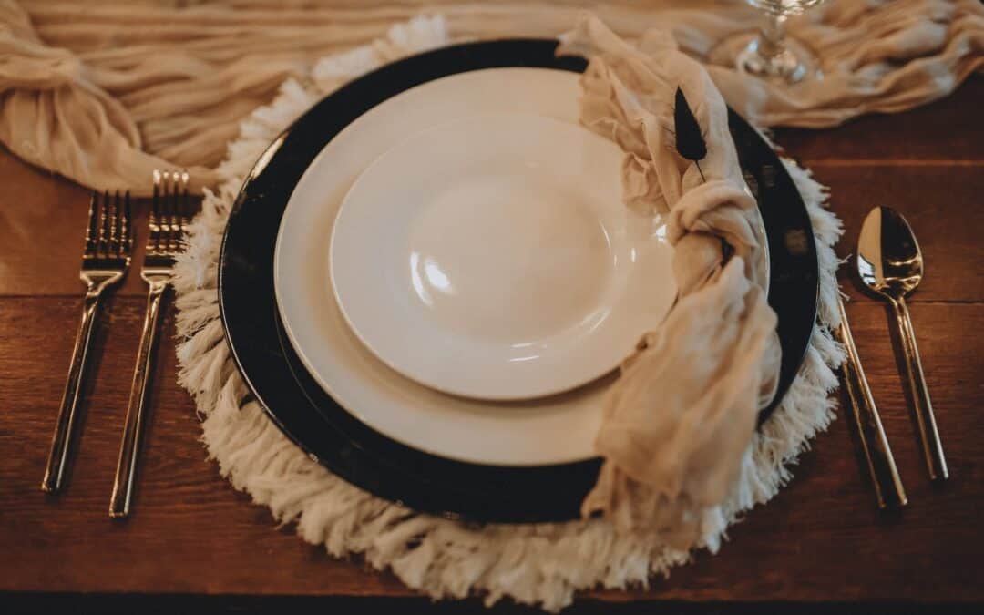 Elegant place setting with layered plates, fringed placemat, silverware, and a fabric napkin on a wooden table.