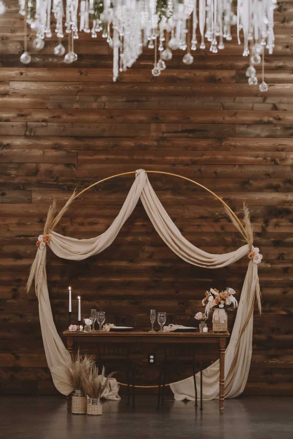 A rustic wedding table with a draped arch, candles, and floral arrangements on a wooden backdrop.