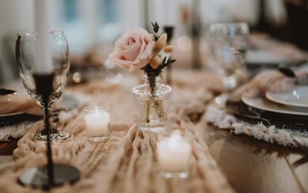 Table set with glassware, a pink rose in a vase, and lit candles on a beige cloth runner. Plates and napkins are arranged in the background.