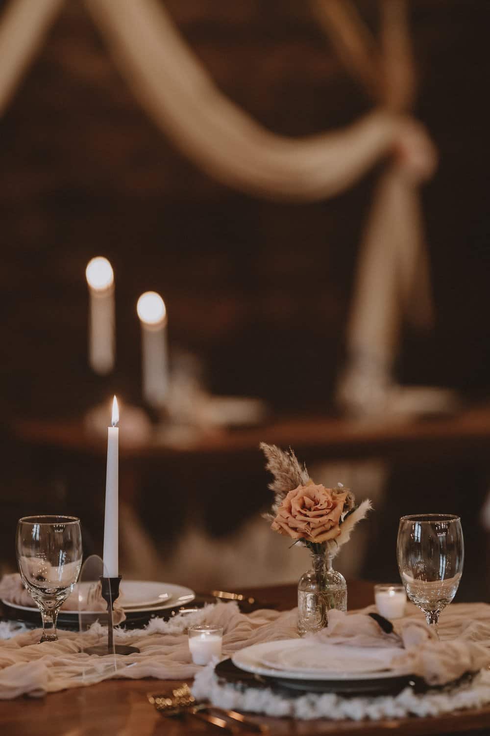 A romantic table setting with a lit candle, wine glasses, plates, and a small floral centerpiece, featuring soft lighting and draped fabric in the background.