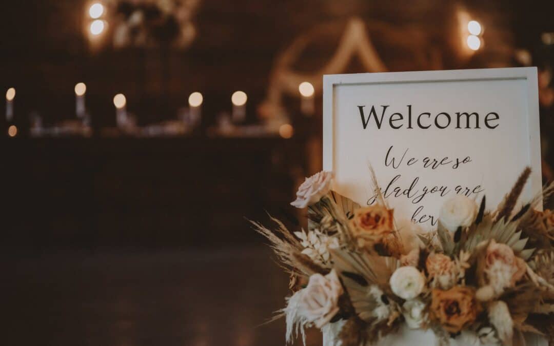 A welcome sign with "We are so glad you are here" text, surrounded by a floral arrangement, in a warmly lit indoor setting.