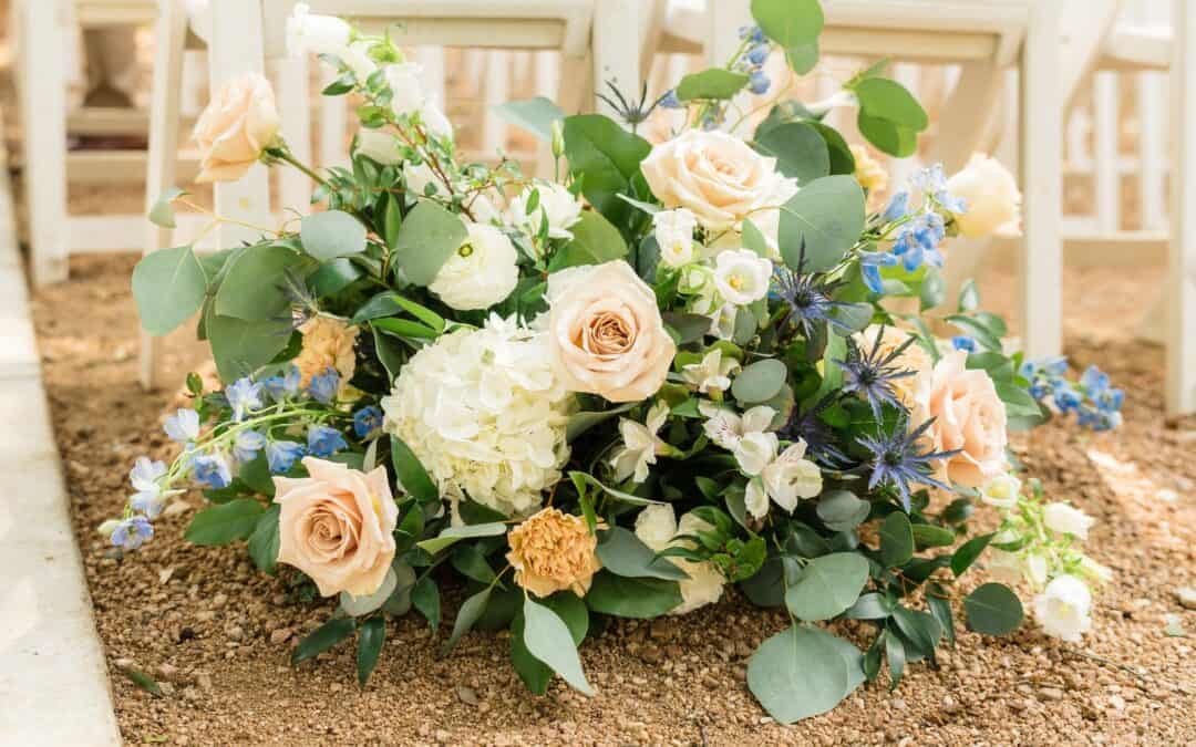 A lush floral arrangement with white hydrangeas, pastel roses, and blue flowers sits on a gravel path next to white wooden chairs.