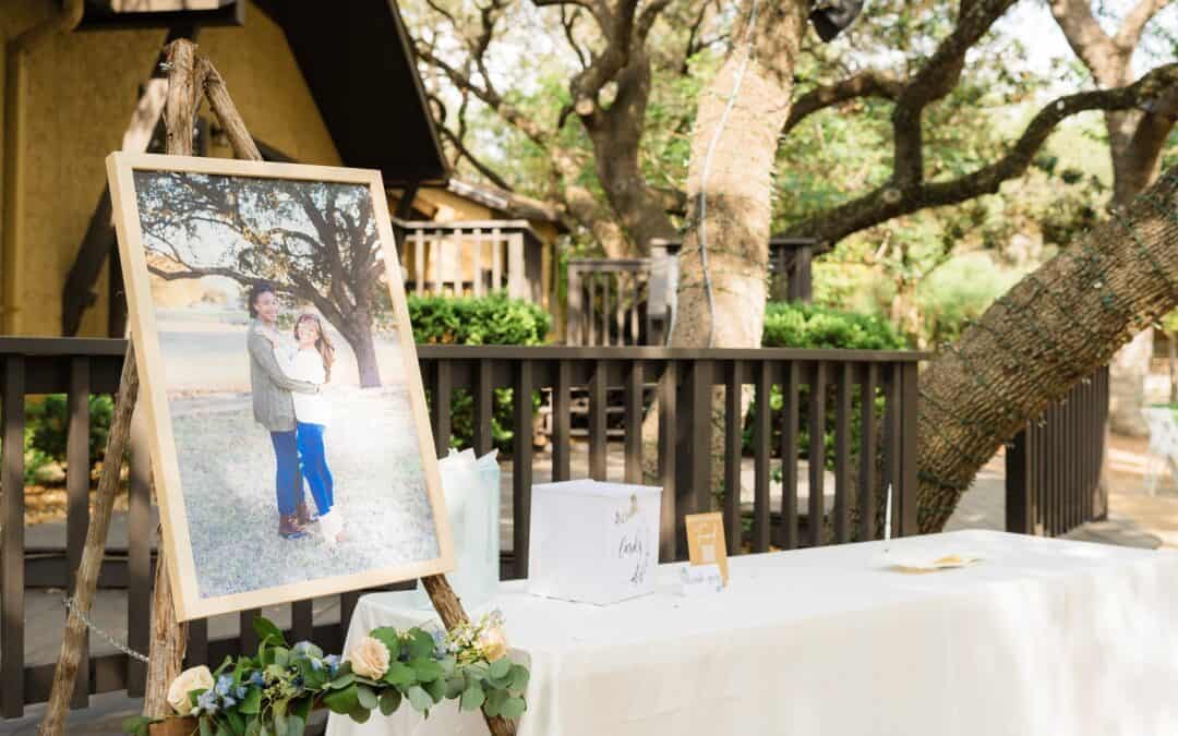 Framed couple's photo on an easel next to a table set up outdoors with a card box and small floral arrangement.