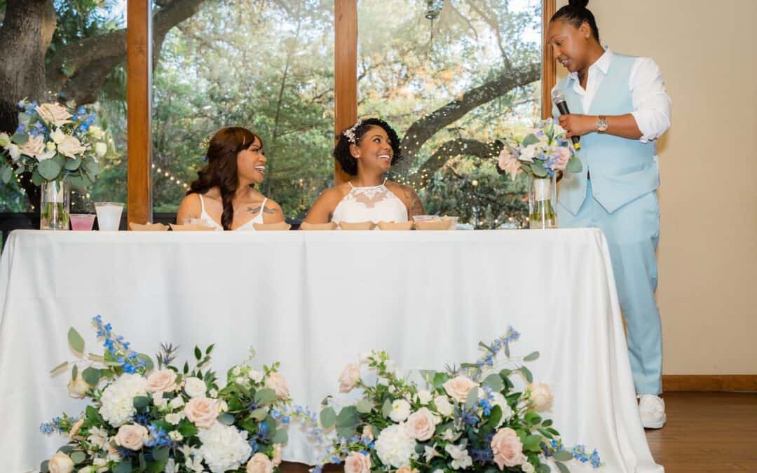 Three people at a decorated table with flowers: two seated in wedding attire smiling, and one in a light blue suit speaking into a microphone.