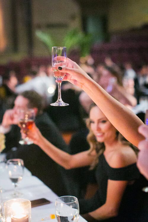 People raising glasses in a toast at a formal event, with a candlelit table in the foreground.